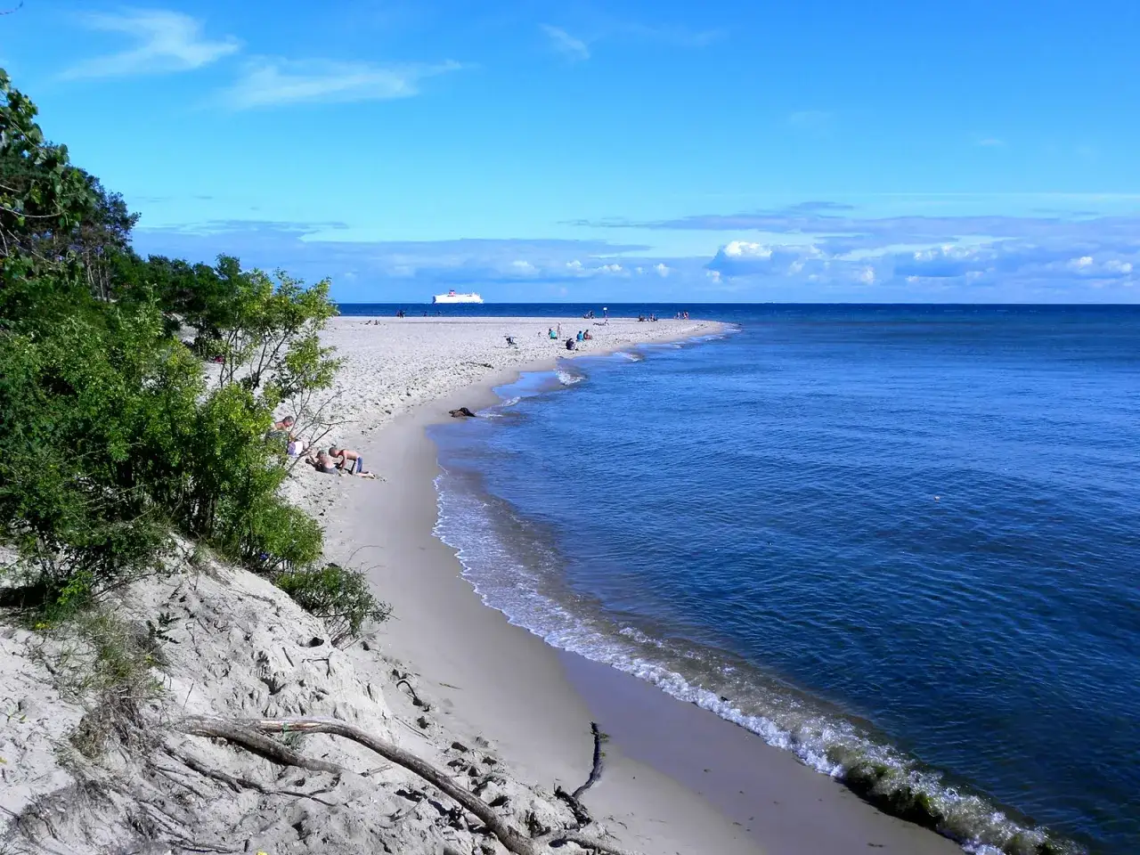 Słoneczny dzień na piaszczystej plaży, z zielonymi wydmami i błękitnym morzem. W oddali widać statek, a na brzegu odpoczywają ludzie. Typowe polskie wybrzeże miejscowości.