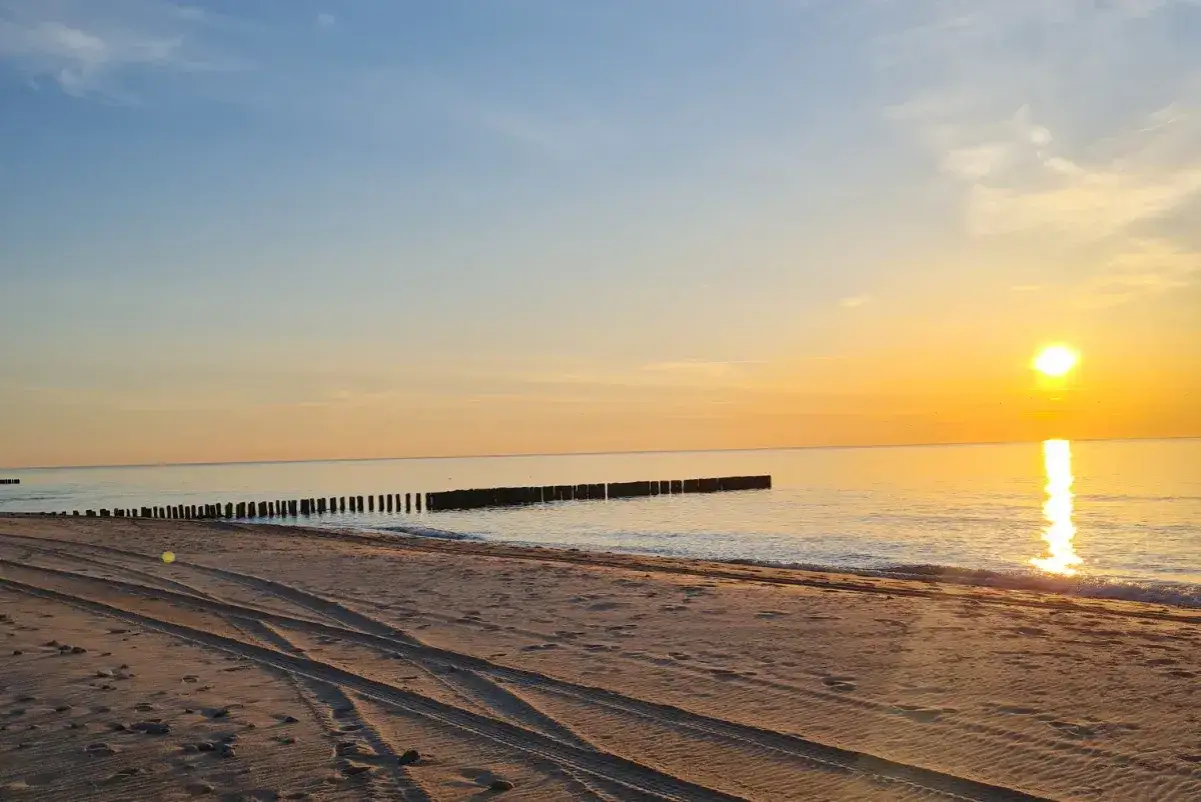 Zachód słońca nad spokojnym morzem, złocący piaszczystą plażę z widocznymi śladami opon. W oddali widać falochrony. Idealne wicie nad morzem.