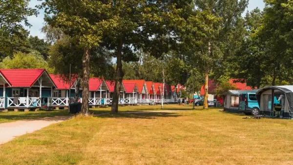 Rząd kolorowych domków letniskowych w Łebie z czerwonymi dachami, otoczonych zielenią. Obok namioty kempingowe i kamper.