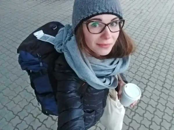 Kobieta w czapce i szaliku trzyma kubek kawy, obok plecak.