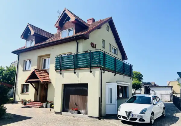 Tanie kwatery prywatne nad morzem. Dom z balkonem i białym samochodem przed posesją, otoczony zielenią.